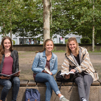 Drei Studentinnen sitzen auf einer Bank, halten Bücher in den Händen und lächeln.
