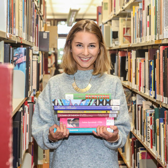 Studierende steht mit Büchern im Arm zwischen den Bücherregalen in der Bibliothek.