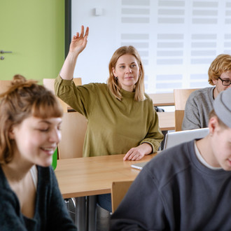 Studierende im Seminarraum sitzen an den Tischen. Eine Person meldet sich. 