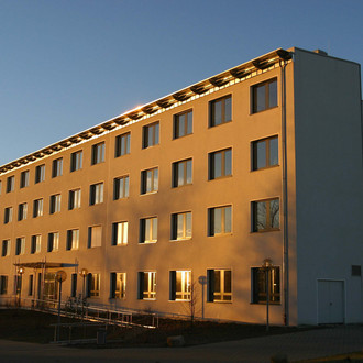 Gebäude E-Technik: Ein hohes weißes Gebäude, das von der Sonne angestrahlt wird. Vor dem Gebäude eine Rampe und über dem Eingang ein Vordach.