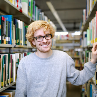 Studierender steht zwischen den Bücherregalen in der Bibliothek.