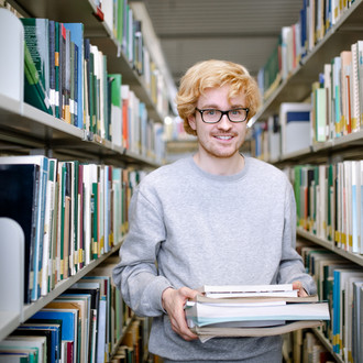 Student zwischen Bücherregalen mit Büchern in der Hand.