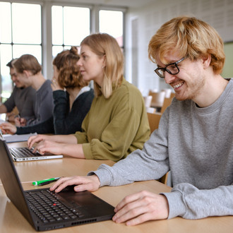 Studierende im Seminarraum sitzen in einer Tischreihe vor ihren Laptops. 