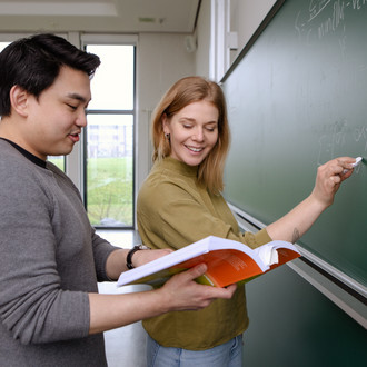 Zwei Studierende an der Tafel, er hält Buch, sie schaut rein, beide lächeln