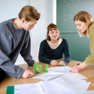 Studierende arbeiten zu dritt an einem Tisch, auf dem das TU-Logo steht.