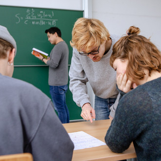 Drei Studierende arbeiten in einem Seminarraum zusammen. Der Dozent ist zur Tafel gewandt.