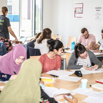 Mehrere Personen sitzen an Tischen und haben Unterlagen vor sich. Eine Person steht und trägt ein T-Shirt mit der Aufschrift "HelpDesk-Mathe".