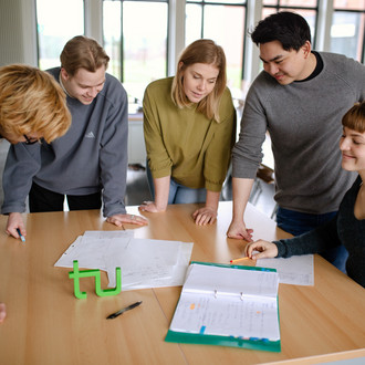 Fünf Studierende im Seminarraum stehen um einen Tisch herum und sehen auf ein Dokument