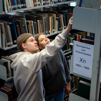 Zwei Studierende suchen nach Büchern in einem Bücherregal in der Bibliothek.