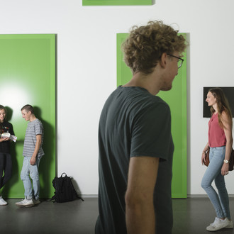 Zwei Studierende lehnen an der Wand, zwei kommen von rechts ins Bild und einer geht nach rechts.