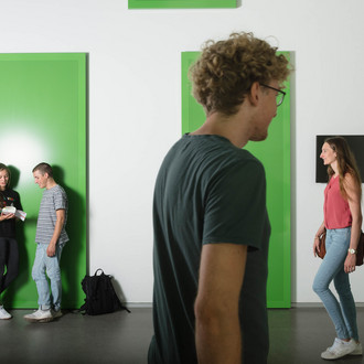 Zwei Studierende lehnen an der Wand, zwei kommen von rechts ins Bild und einer geht nach rechts.