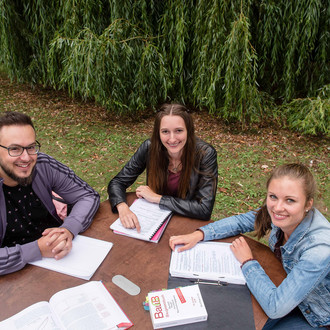 Drei Studierende sitzen an einem Tisch auf einer Wiese und lernen.