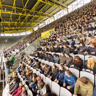 Studierende des ersten Semesters sitzen bei der Erstsemesterbegrüßung auf den Rängen im Stadium und schauen seitlich einem Showact zu.